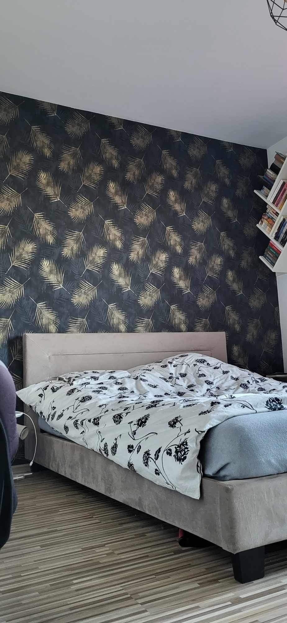 Sypialnia z łóżkiem kontynentalnym, ściana z tapetą w ciemne liście, biała pościel w czarne kwiaty i regał z książkami w rogu.