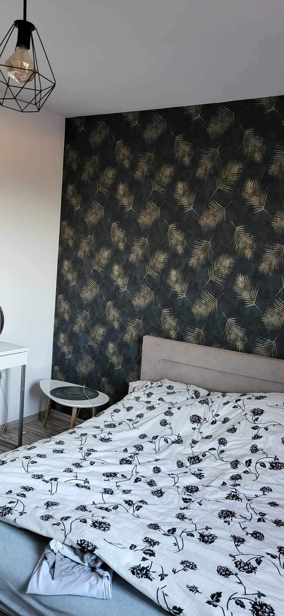 Sypialnia z tapetą w ciemne liście palmowe, białą pościelą w czarne kwiaty, stolikiem pomocniczym i lampą wiszącą w stylu industrialnym.