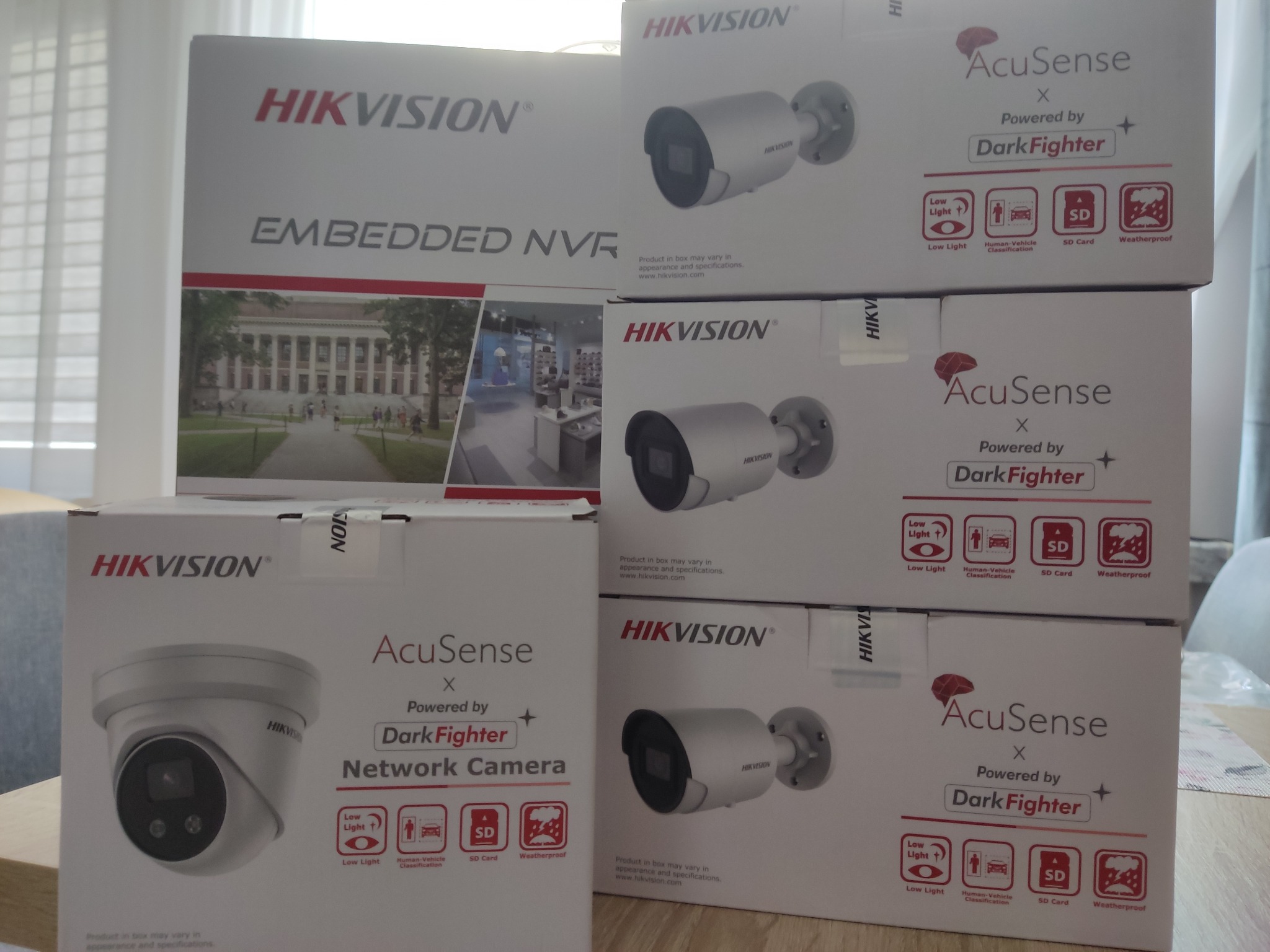 Stos pudełek z kamerami i rejestratorem Hikvision AcuSense DarkFighter, prezentujących specyfikacje: detekcja ludzi/pojazdów, nagrywanie na SD, odporność na warunki atmosferyczne. Dodatkowo pudełko...
