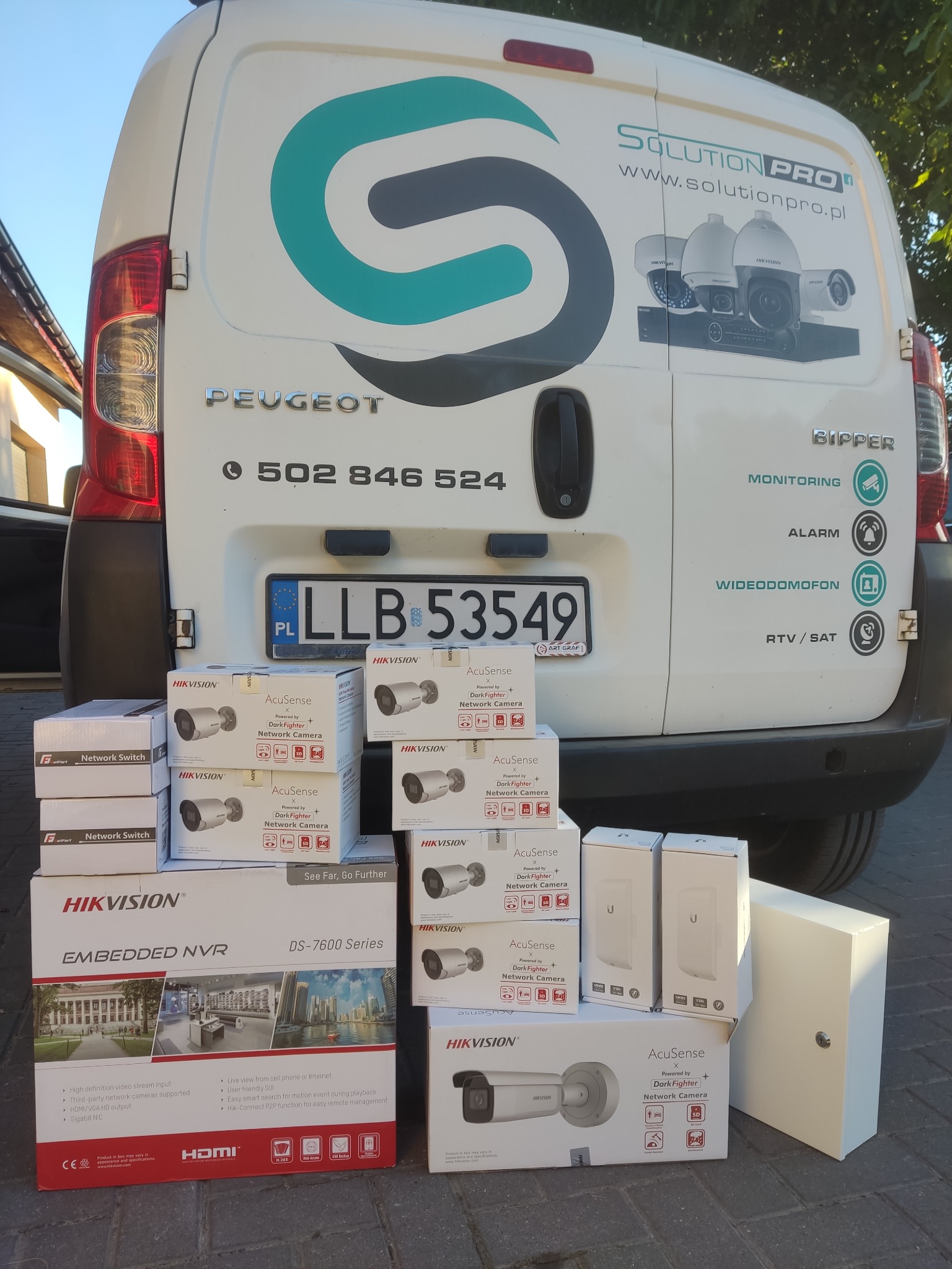 Pudełka z kamerami sieciowymi Hikvision AcuSense i rejestratorem NVR DS-7600 Series na tle białego samochodu dostawczego Peugeot Bipper z reklamą firmy Solution Pro oferującej monitoring, alarmy...