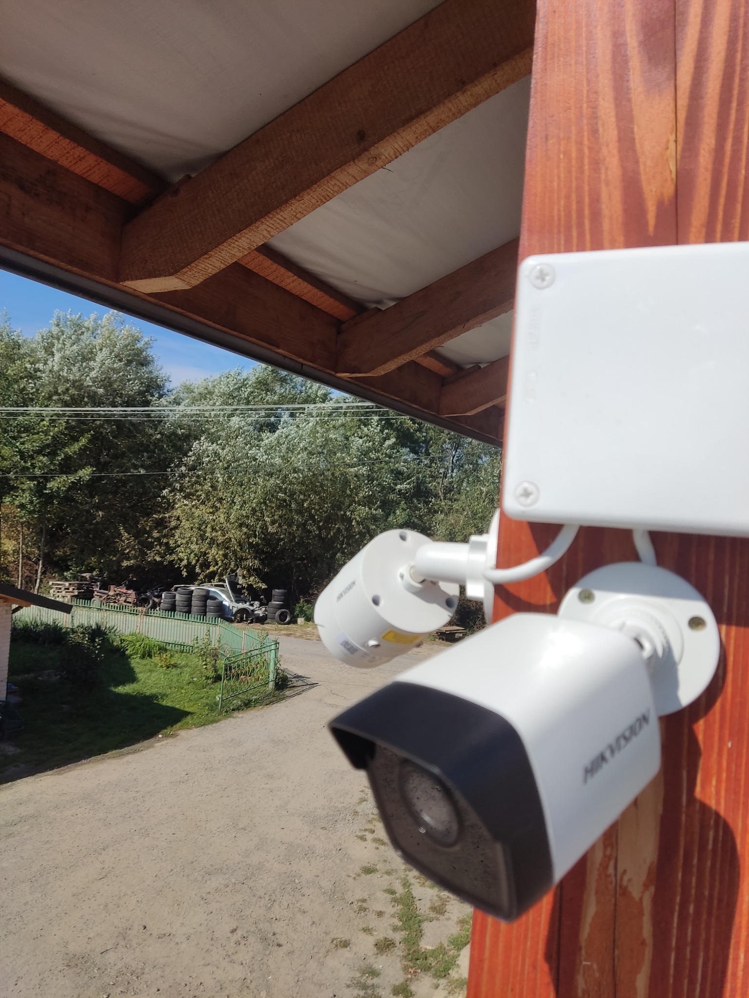 Dwie kamery monitoringu Hikvision zamontowane na drewnianym słupku pod zadaszeniem, widok na podwórze z oponami i drzewami w tle.