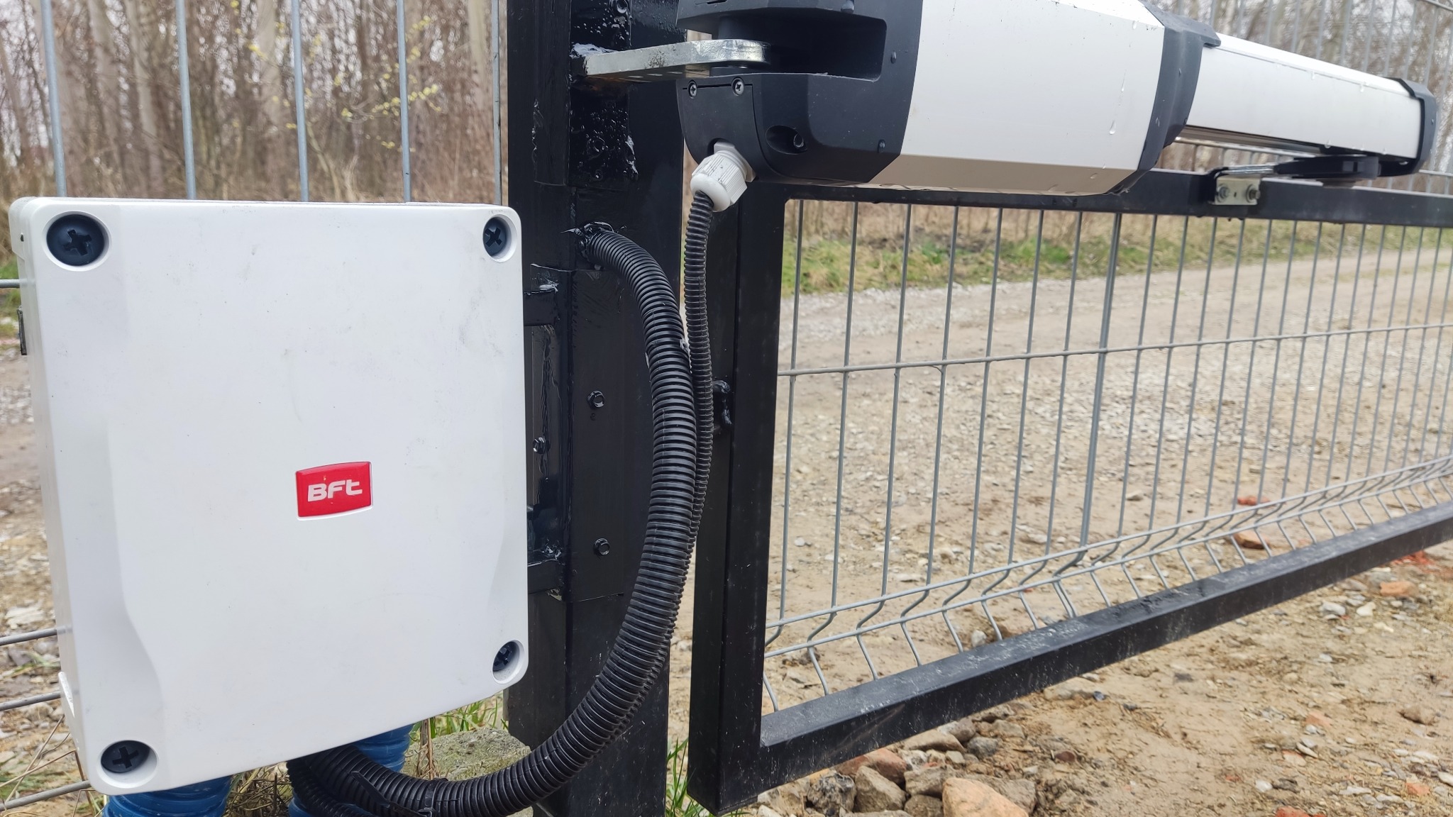 Automat do bramy przesuwnej firmy BFT, z widoczną centralą sterującą w białej obudowie z logo, zamontowany na czarnym słupku bramy wjazdowej.