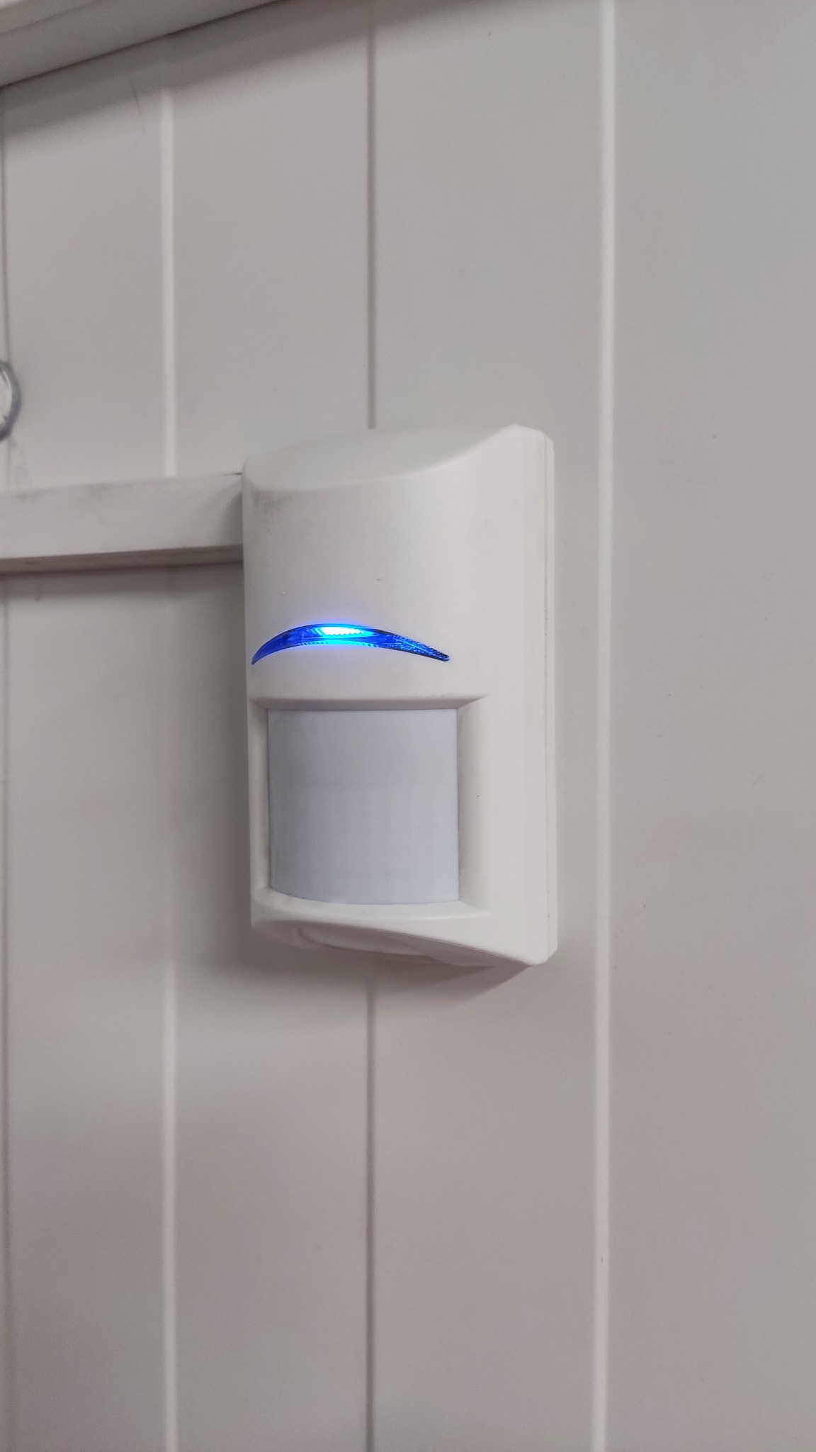 Biały czujnik ruchu z niebieską diodą LED, zamontowany na białej, panelowej ścianie. Widoczne ślady kurzu na obudowie.