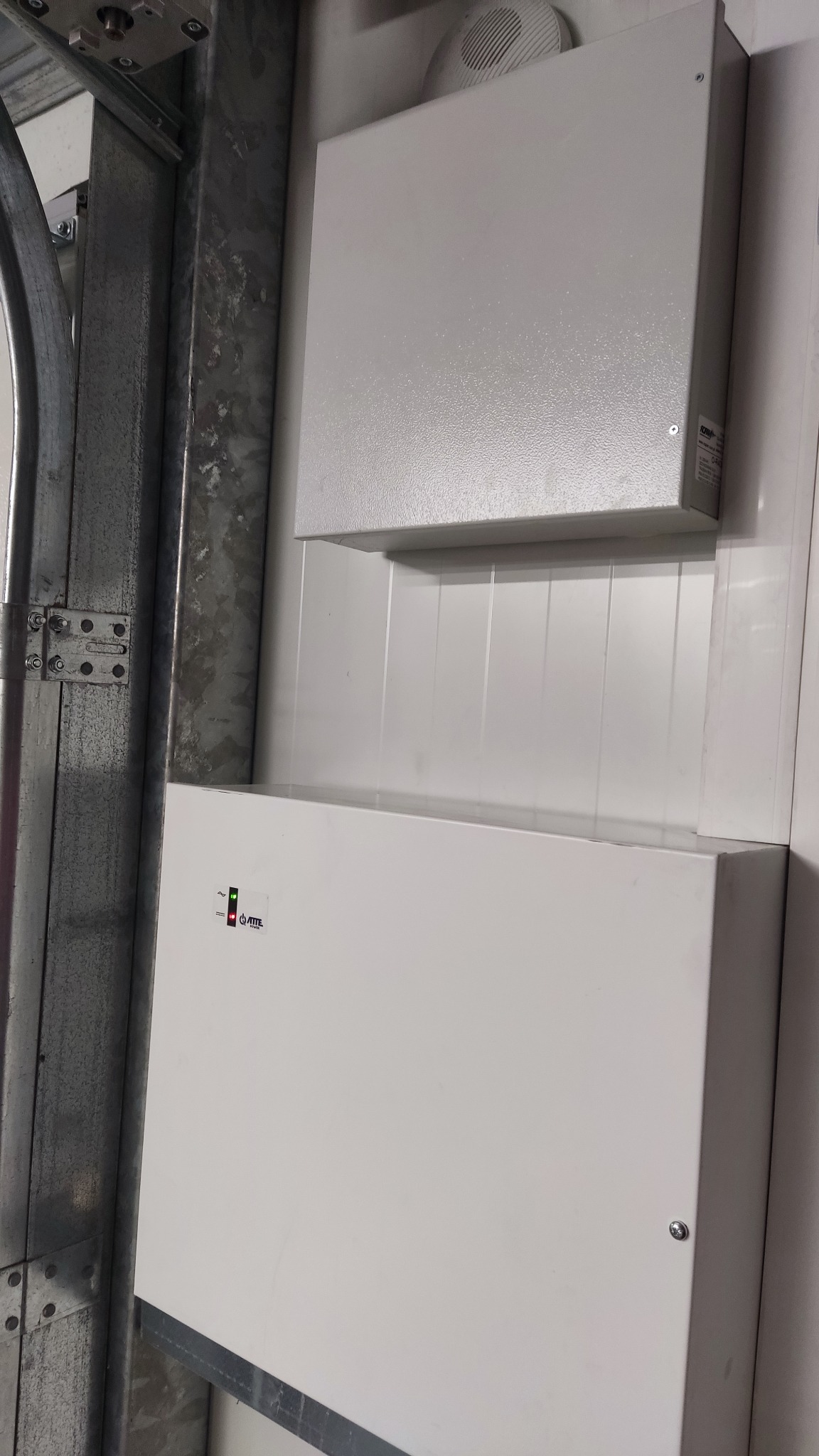 Dwa białe metalowe panele elektryczne zamontowane na ścianie, widoczny alarmowy panel sterujący z diodami LED i naklejką producenta, obok ramy garażowej.
