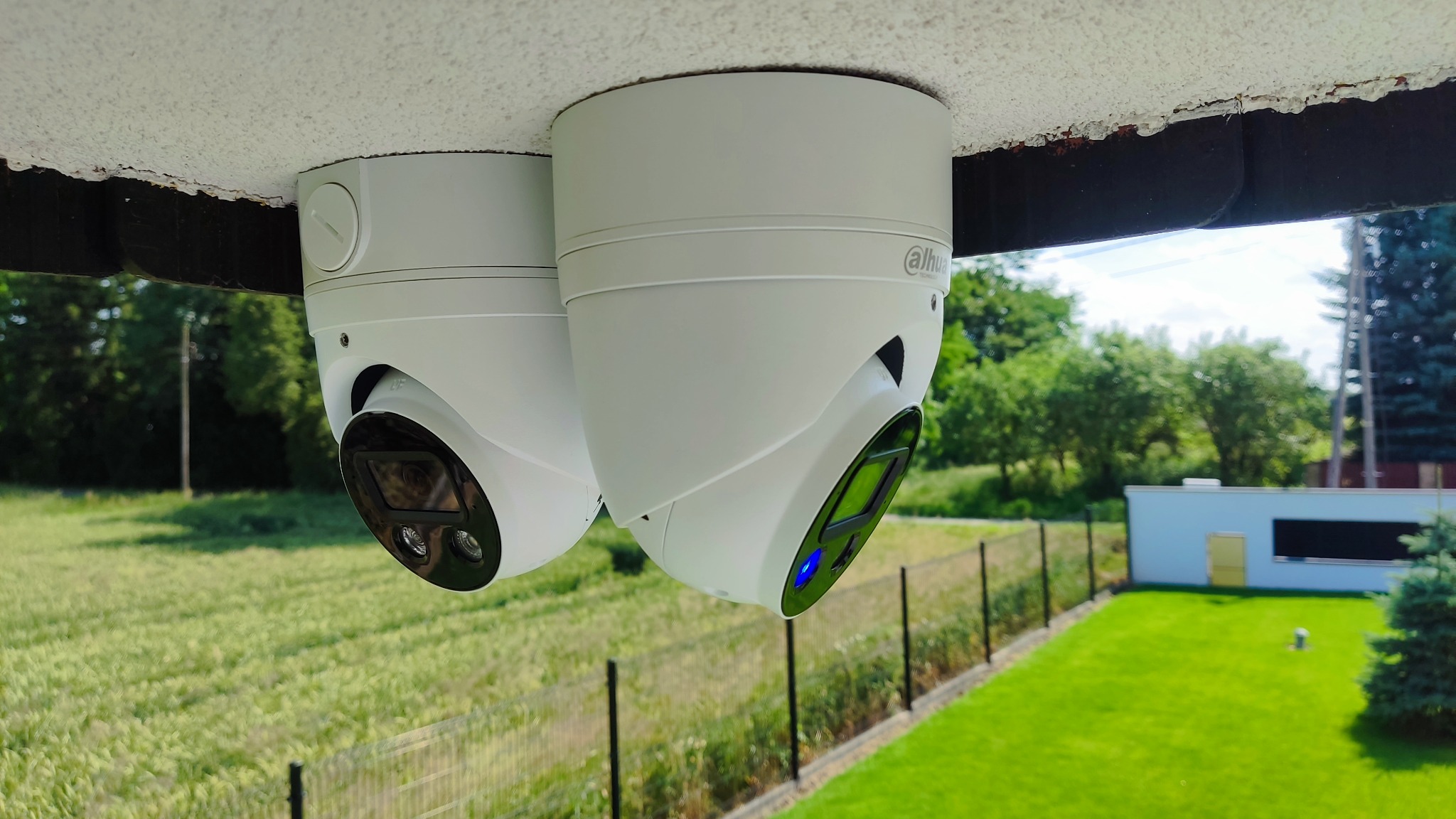 Dwie białe kamery monitoringu Dahua zamontowane pod sufitem z widokiem na zielone podwórko i budynek gospodarczy.