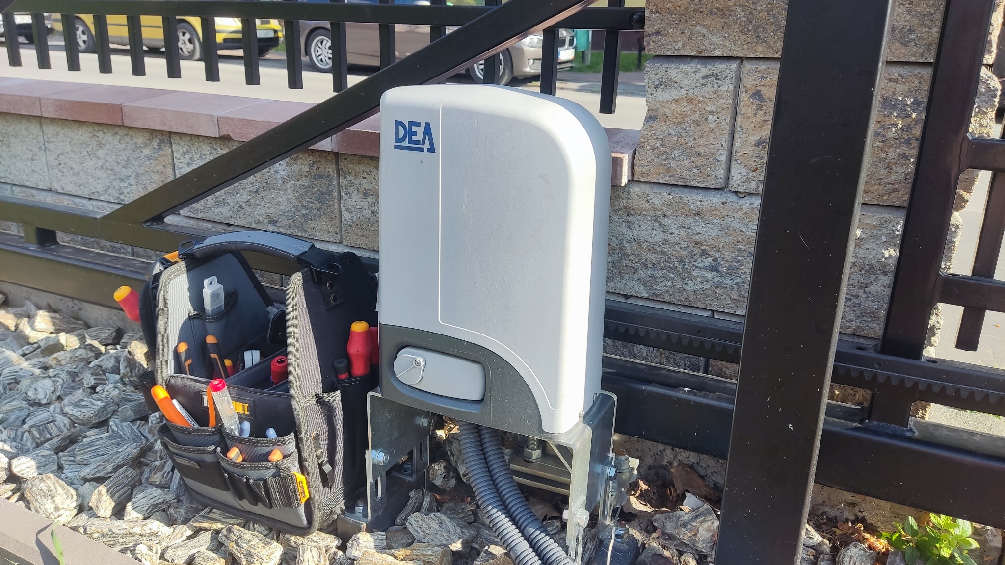 Szary automat do bramy z niebieskim logo DEA, zamontowany na metalowej konstrukcji, obok torba narzędziowa z widocznymi śrubokrętami na tle kamiennej ściany i fragmentu ogrodzenia.