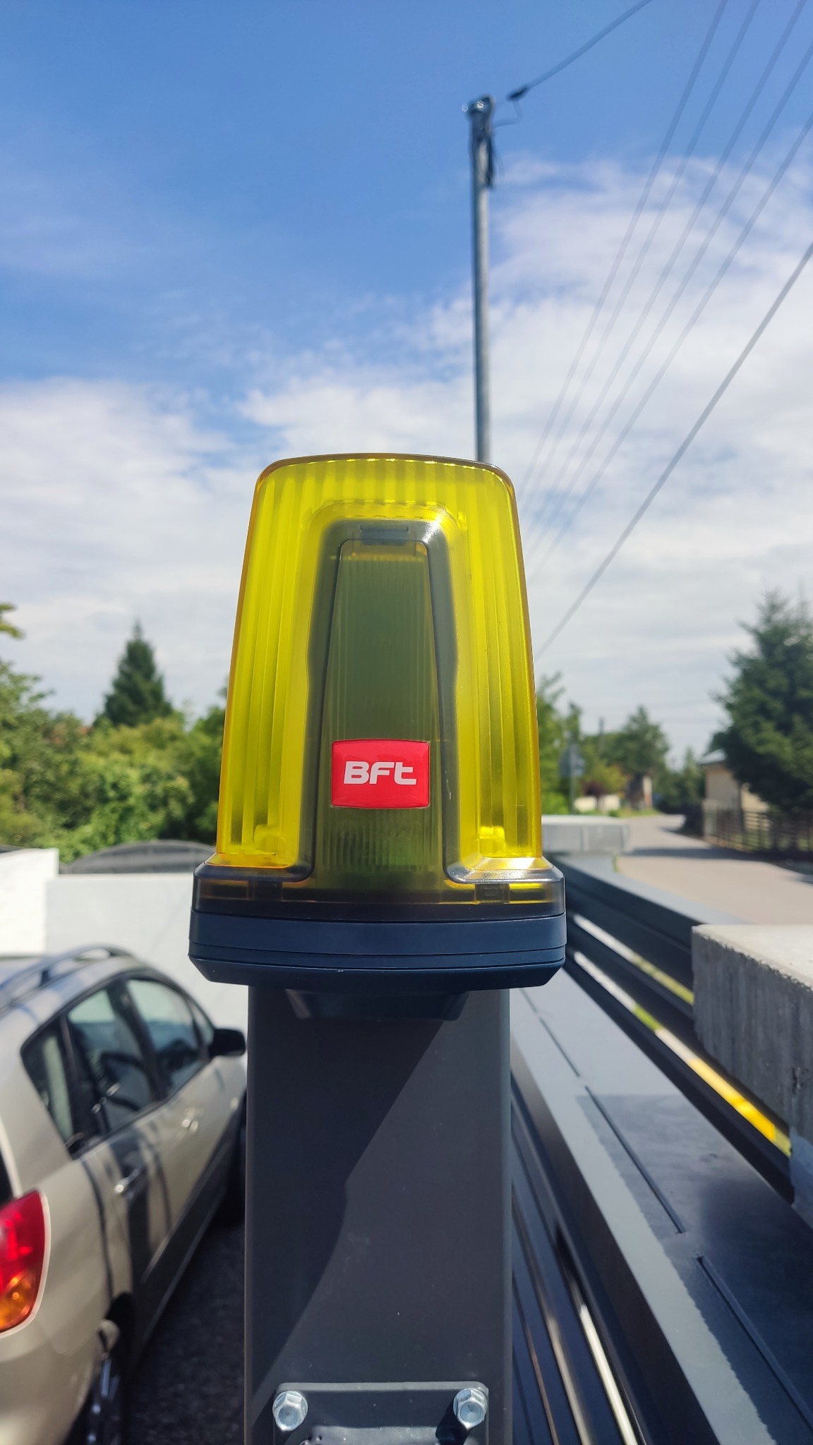 Żółta lampa ostrzegawcza BFT zamontowana na słupku bramy wjazdowej, widok z dołu na tle błękitnego nieba i zieleni drzew.