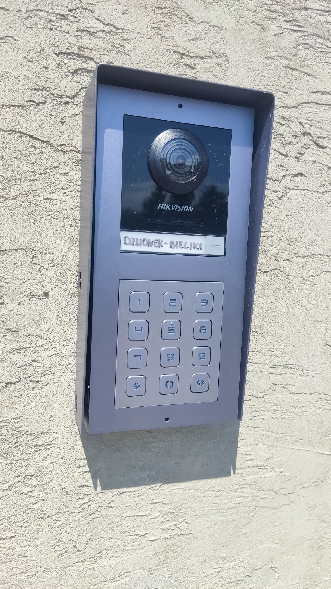 Szary panel wideodomofonu Hikvision z klawiaturą numeryczną i kamerą, zamontowany na teksturowanej, beżowej ścianie zewnętrznej budynku.