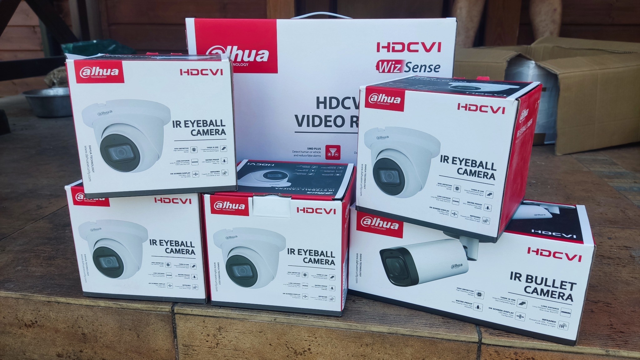 Stos pudełek z kamerami Dahua HDCVI, w tym modele 'IR Eyeball Camera' i 'IR Bullet Camera', na drewnianym podłożu, z większym pudełkiem rejestratora w tle.