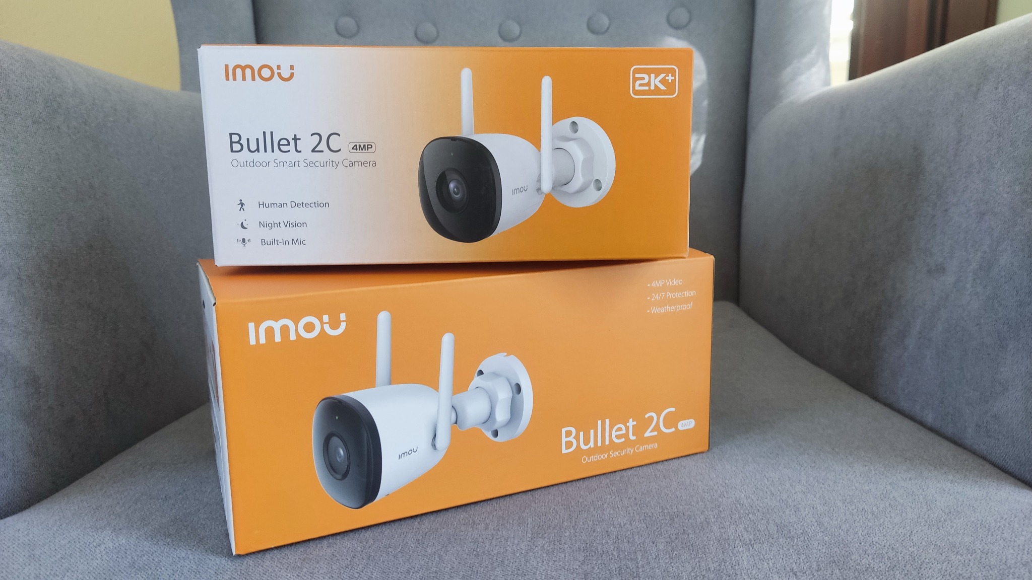 Dwa pudełka kamer IP Imou Bullet 2C 4MP na szarym fotelu, prezentujące produkt w opakowaniu z widocznymi specyfikacjami i cechami, takimi jak detekcja ludzi, tryb nocny i wbudowany mikrofon.