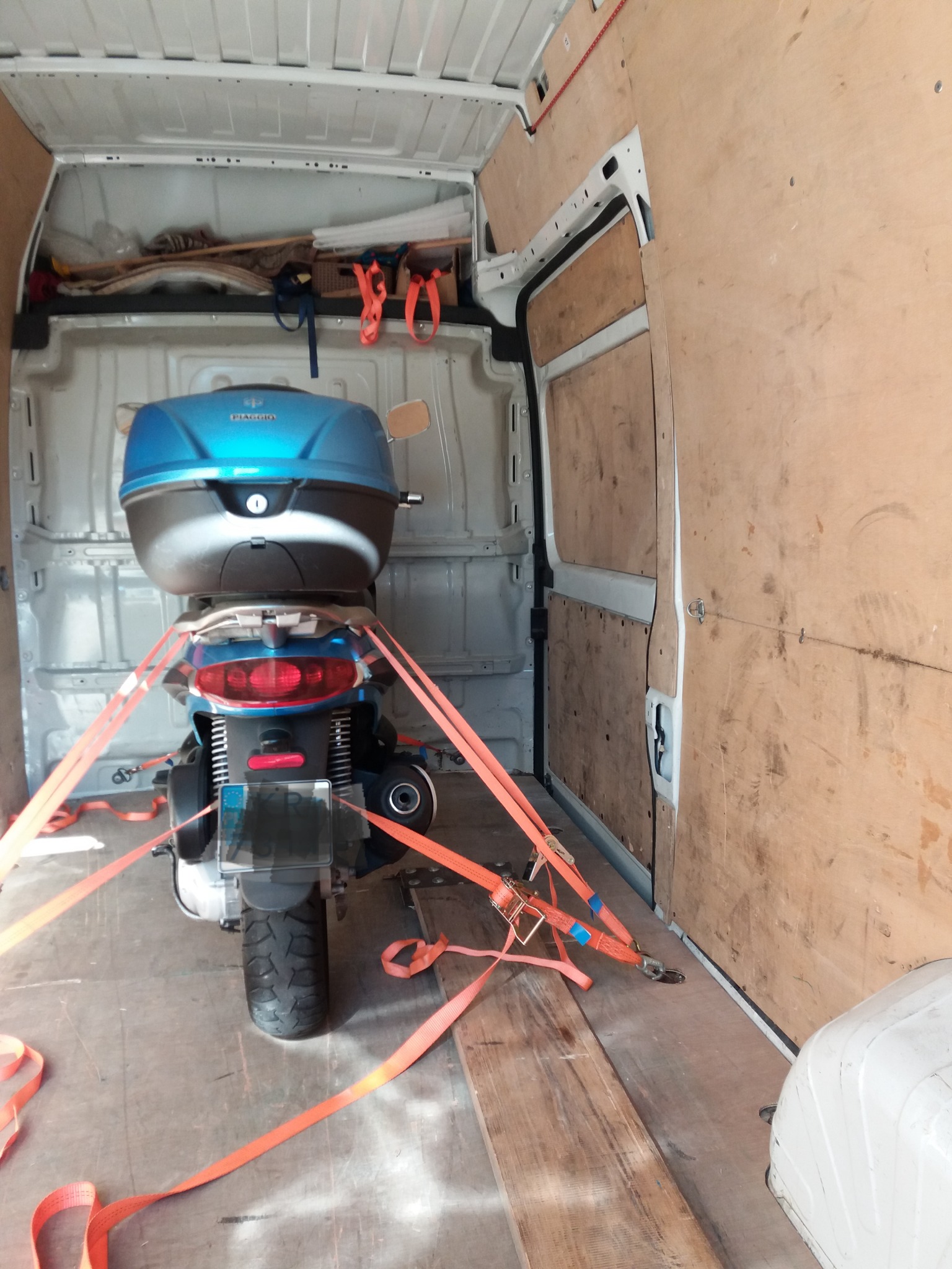 Skuter Piaggio z niebieskim kufrem, zabezpieczony pomarańczowymi pasami transportowymi wewnątrz furgonetki z drewnianą zabudową.