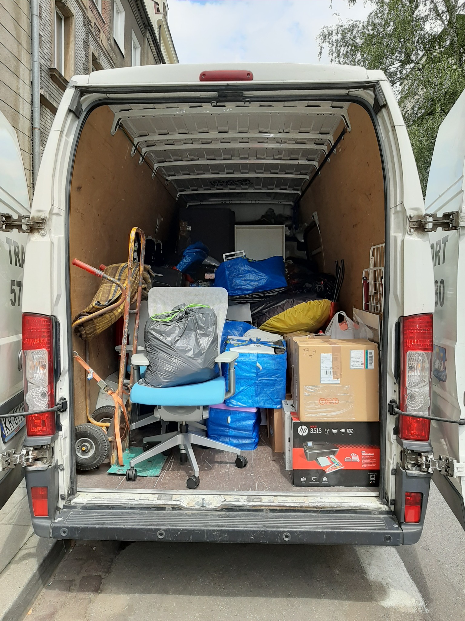 Wnętrze białego busa załadowane meblami, kartonami i workami, widok z tyłu, otwarte drzwi, w tle budynek mieszkalny.