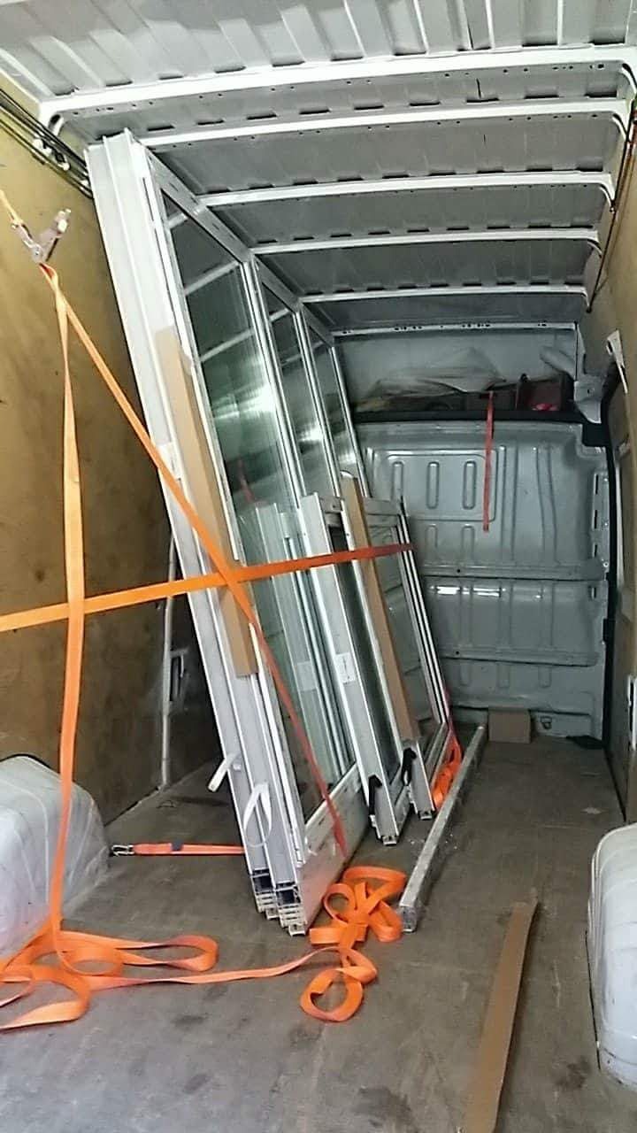Białe okna PCV zabezpieczone pomarańczowymi pasami transportowymi wewnątrz furgonetki podczas transportu.