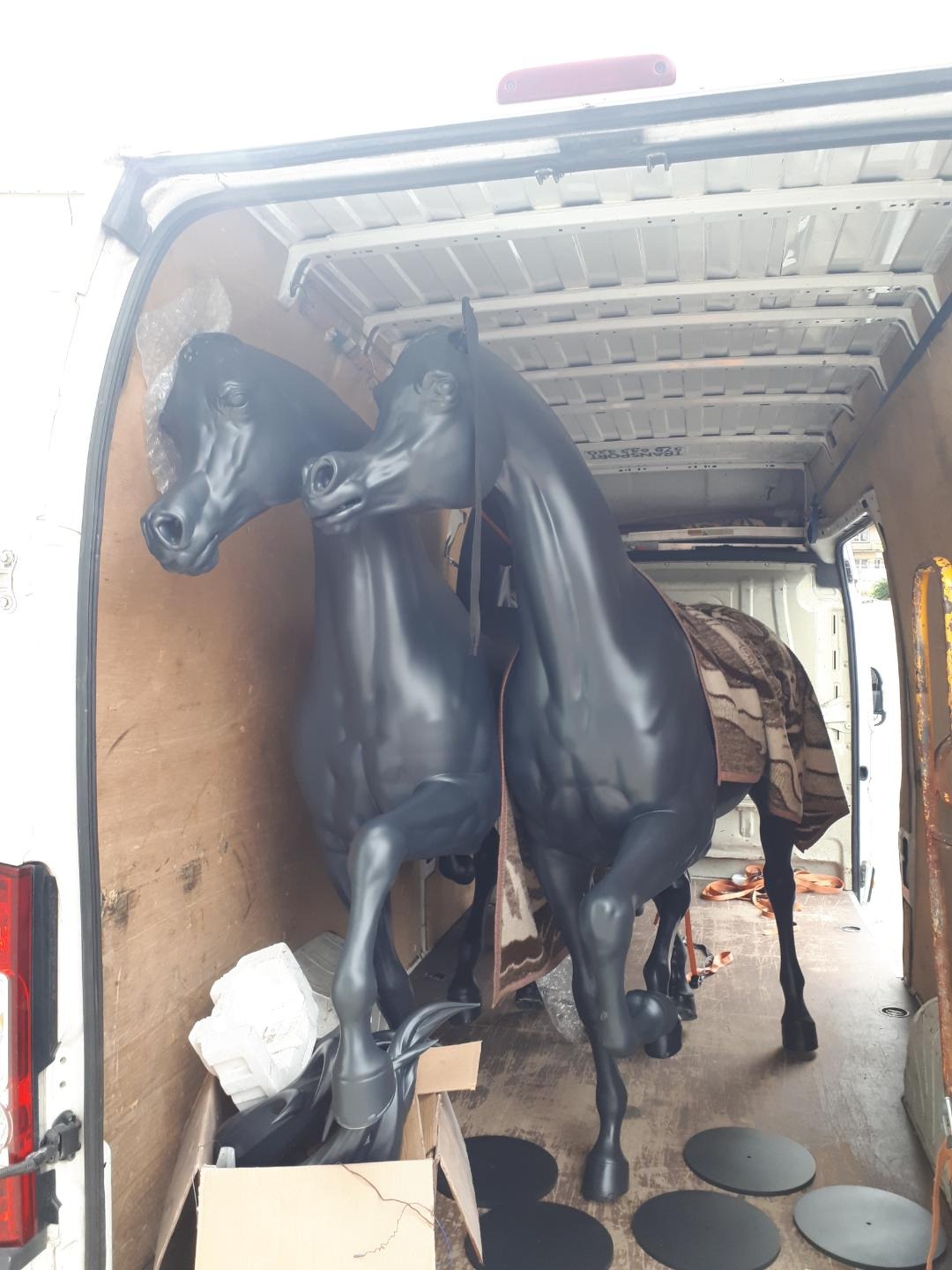 Dwie czarne, błyszczące figury koni przewożone w przestrzeni ładunkowej białego busa, zabezpieczone kocem i kartonami.