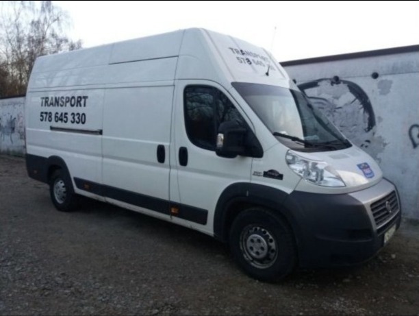 Biały samochód dostawczy z napisem 'TRANSPORT' i numerem telefonu na boku, zaparkowany na żwirowym podłożu przy szarym murze z graffiti.