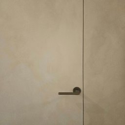 STRUKTURY WROCŁAW SPÓŁKA Z OGRANICZONĄ ODPOWIEDZIALNOŚCIĄ - Beżowe drzwi z ukrytą ościeżnicą i klamką na tle ściany o delikatnej fakturze, wykończonej techniką lime wash.