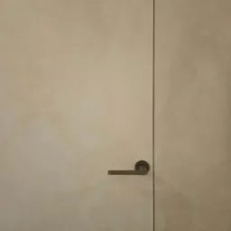 Beżowe drzwi z ukrytą ościeżnicą i klamką na tle ściany o delikatnej fakturze, wykończonej techniką lime wash.