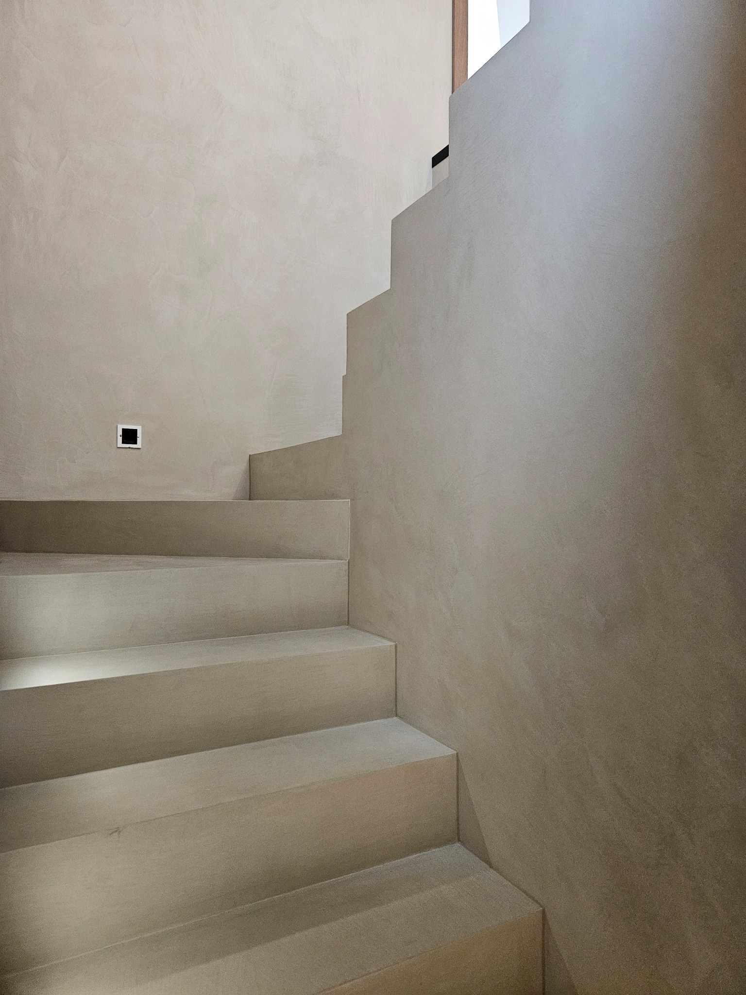 Schody i ściana wykończone w beżowym mikrocemencie, delikatna faktura, minimalistyczne oświetlenie, nowoczesne wnętrze, widok z dołu.