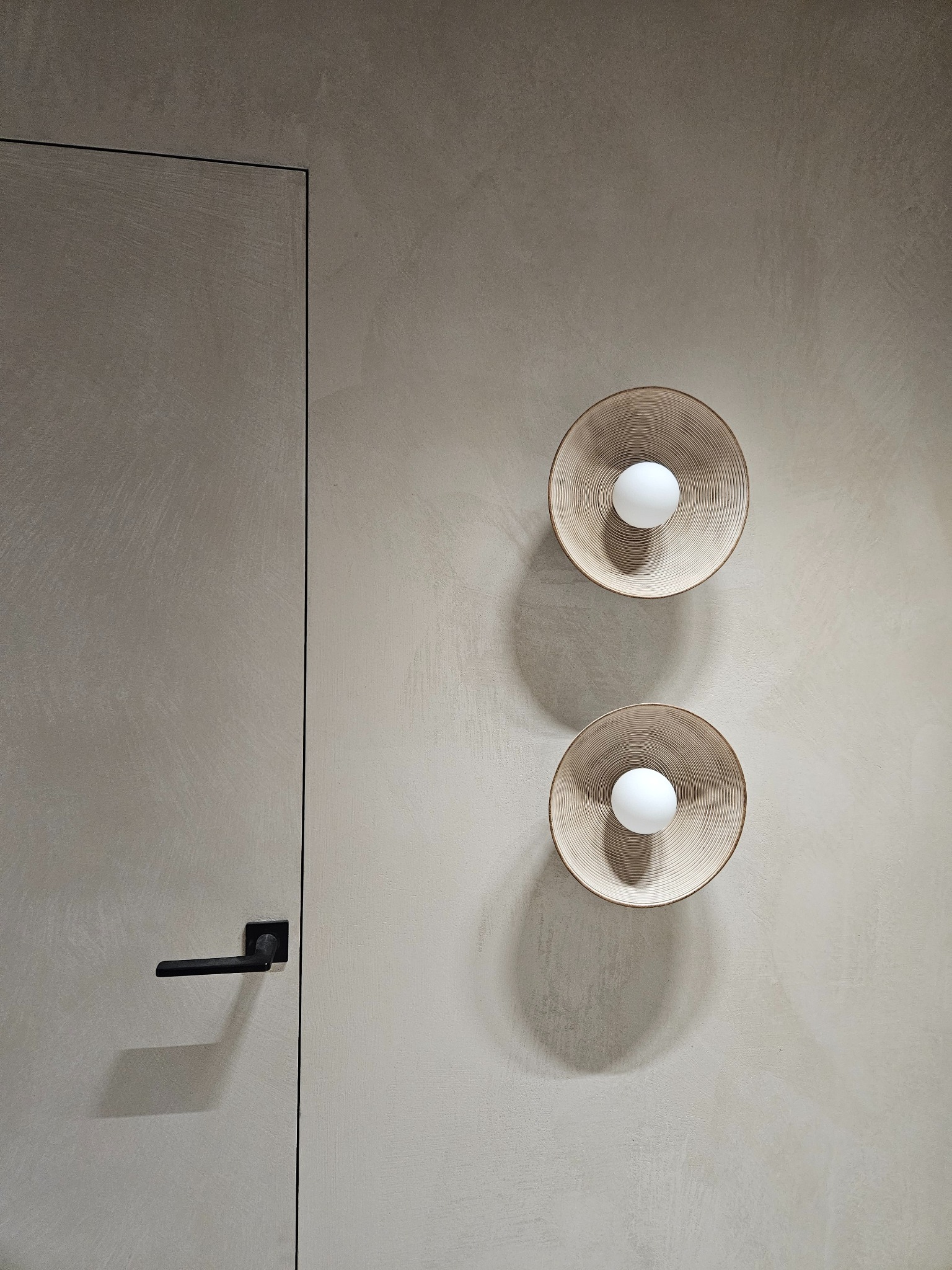 Minimalistyczne wnętrze: drzwi z czarną klamką i ściana pokryta mikrocementem, oświetlona dwoma okrągłymi lampami z żarówkami.