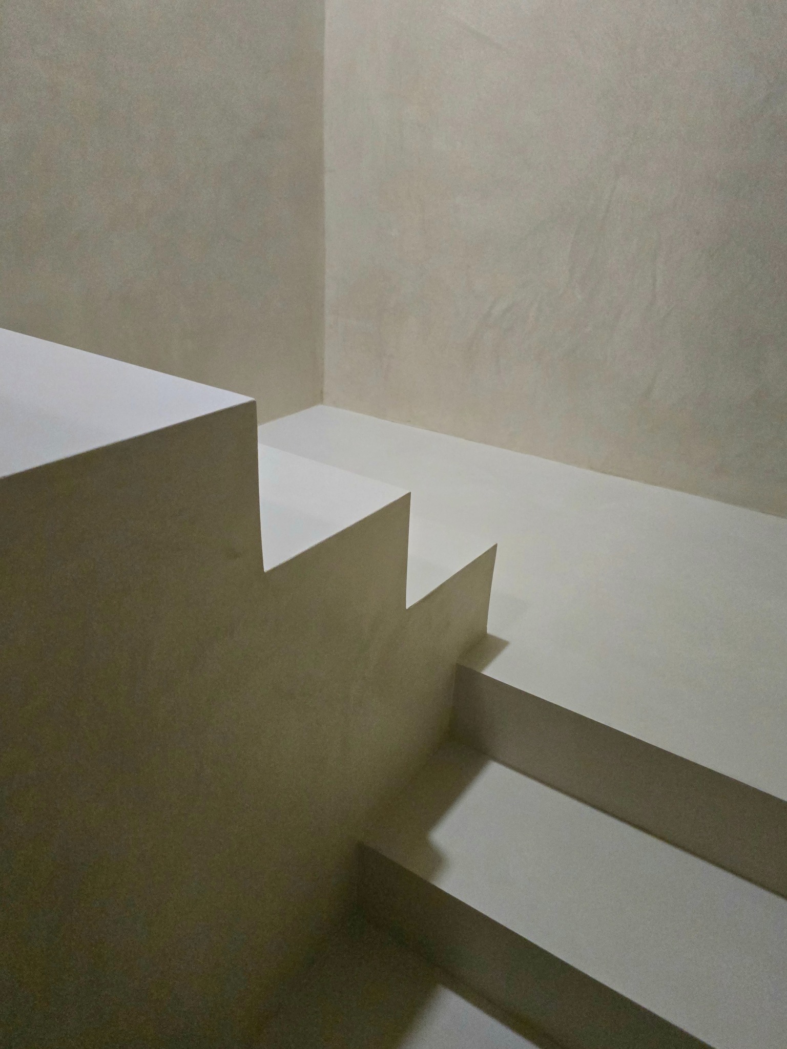Minimalistyczne schody z mikrocementu w odcieniach beżu, z delikatną fakturą na ścianach i schodach. Ujęcie z dołu, podkreślające geometrię i gładkie powierzchnie.