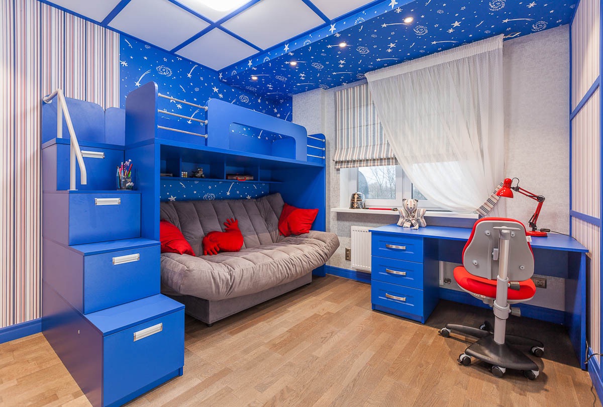 Pokój dziecięcy w niebieskiej tonacji z łóżkiem piętrowym ze schodami pełniącymi funkcję szuflad, rozkładaną sofą na dole, biurkiem z czerwoną lampką i krzesłem na kółkach oraz tapetą w gwiazdy...