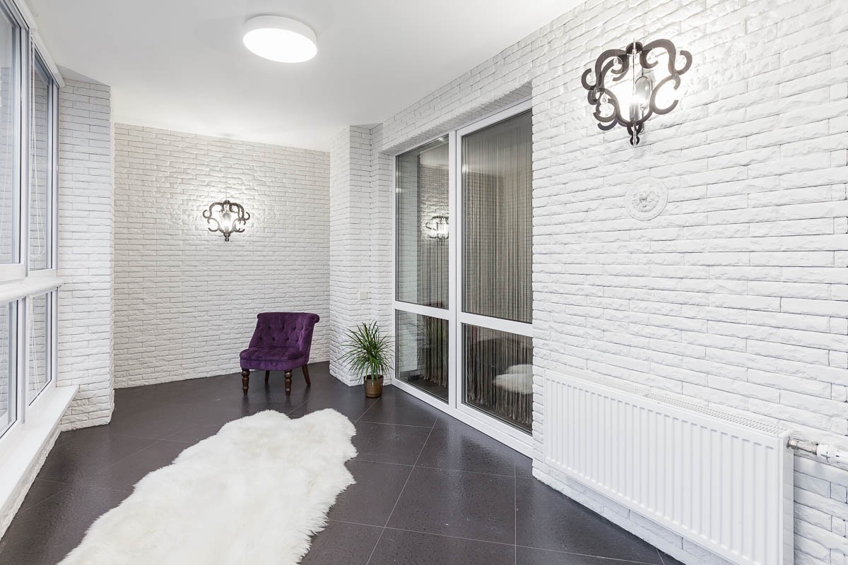 Wnętrze z białej cegły dekoracyjnej, oświetlone ozdobnymi kinkietami, z fioletowym krzesłem i białym dywanem na ciemnej podłodze.