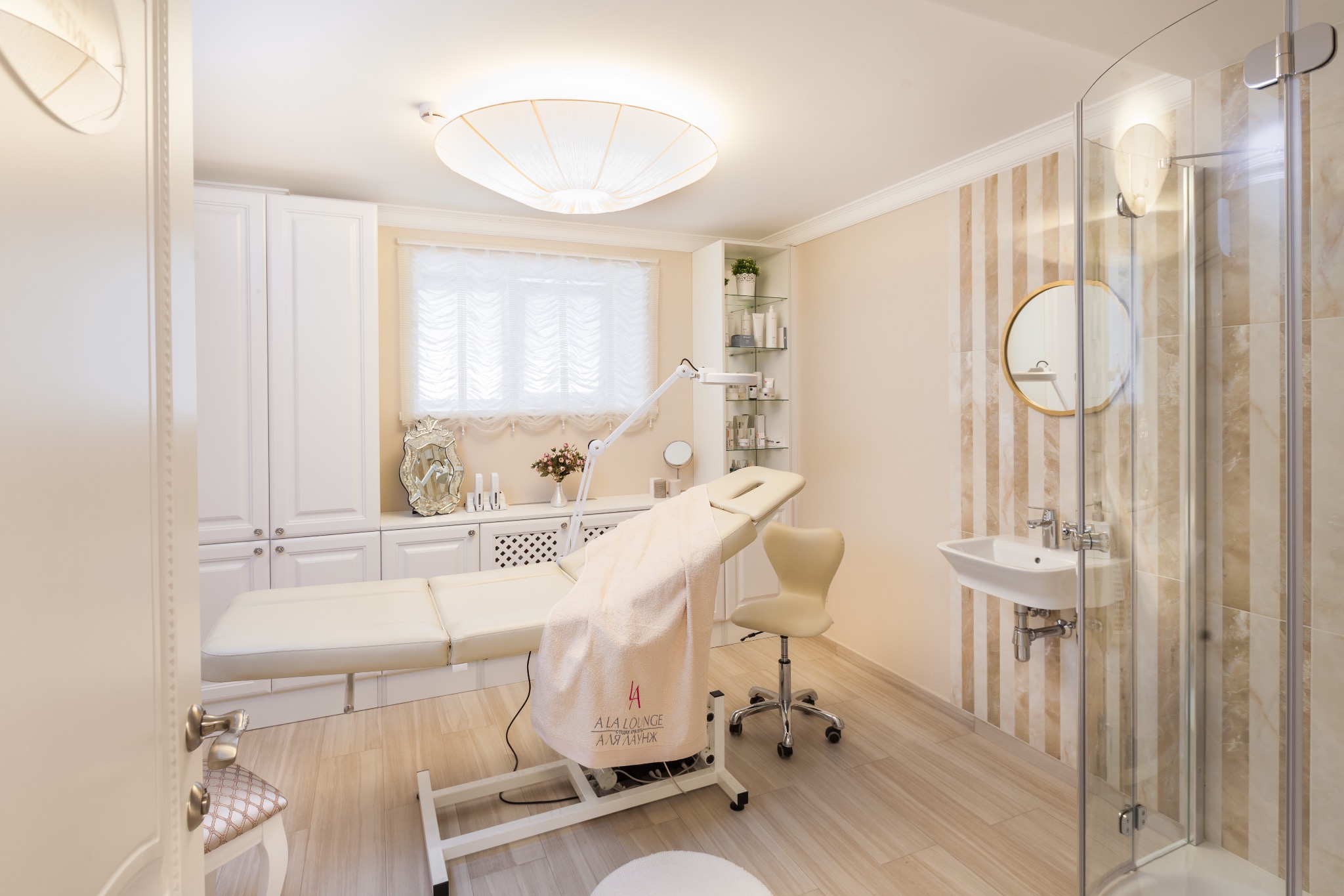 Jasne wnętrze gabinetu kosmetycznego z beżowym łóżkiem do zabiegów, krzesłem na kółkach, umywalką i kabiną prysznicową w tle.