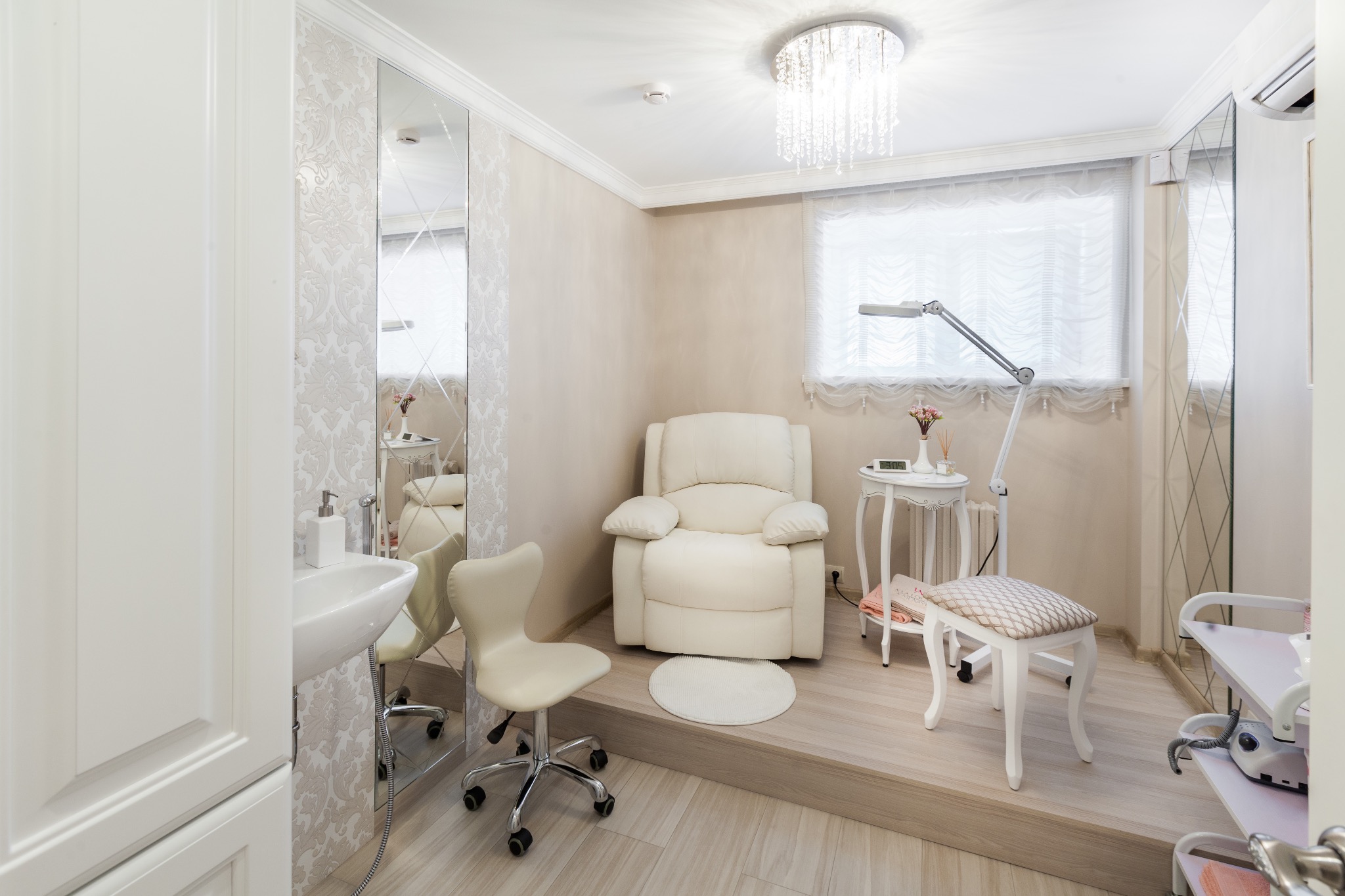 Jasne wnętrze salonu kosmetycznego z fotelem do pedicure, umywalką, lustrem z dekoracyjnymi szlifami i kryształowym żyrandolem.