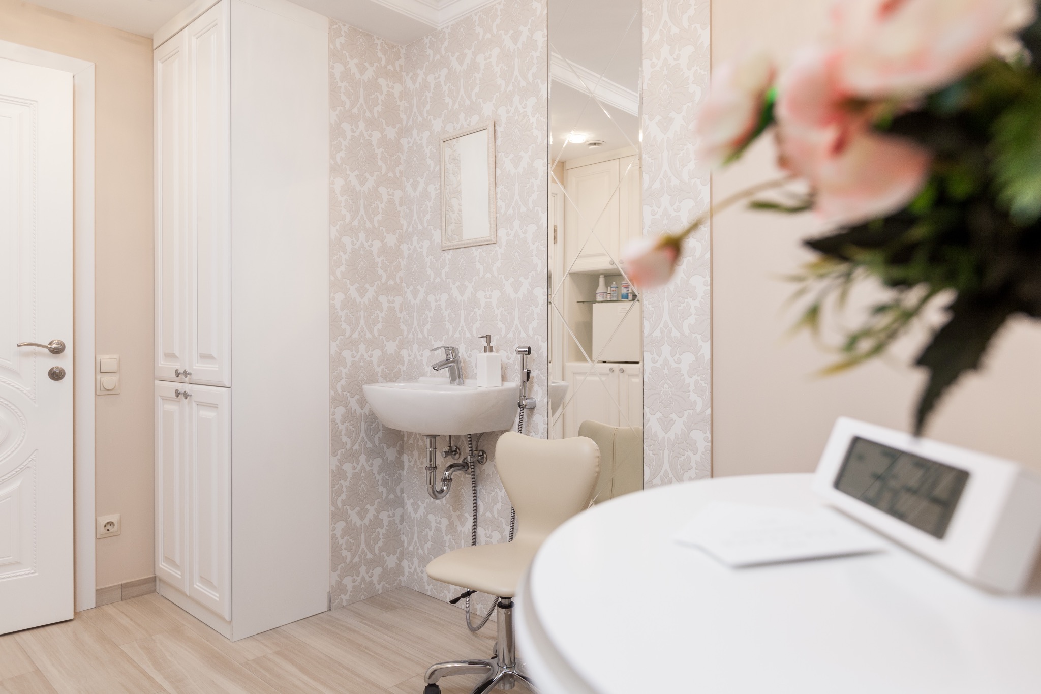 Nowoczesna, jasna łazienka z umywalką nablatową, lustrem w ozdobnej ramie, tapetą w delikatny wzór i białymi meblami, widoczny fragment blatu z elektronicznym zegarem.