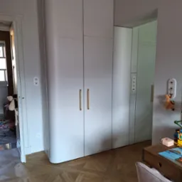 Biała szafa z zaokrąglonym narożnikiem, złotymi uchwytami i panelami dekoracyjnymi, w pokoju dziecięcym z drewnianą podłogą w jodełkę i biurkiem z zabawkami.