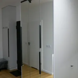 Biała szafa wnękowa z lustrzanymi drzwiami przesuwnymi, widoczna w przedpokoju z drewnianą podłogą i czarnymi akcentami, takimi jak oświetlenie i wieszak.