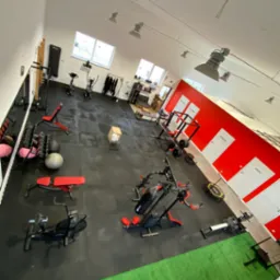 Widok z góry na salę treningową wyposażoną w różnorodny sprzęt, w tym maszyny siłowe, hantle, rowery stacjonarne i ergometry wioślarskie; ściany w kolorze białym i czerwonym, podłoga wyłożona...