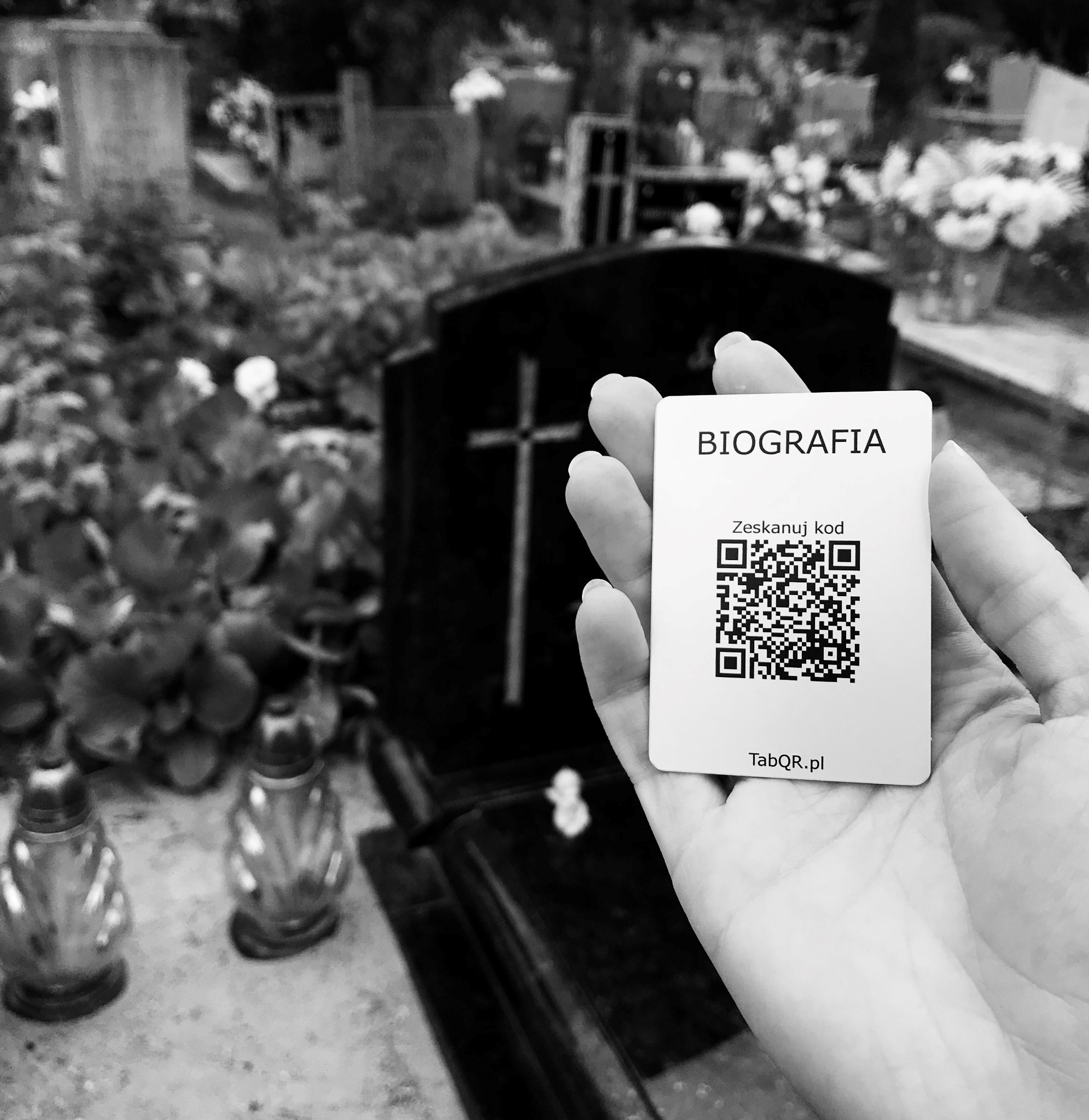 Czarno-białe zdjęcie dłoni trzymającej kartę z kodem QR i napisem 'Biografia' na tle nagrobka z krzyżem i cmentarza.