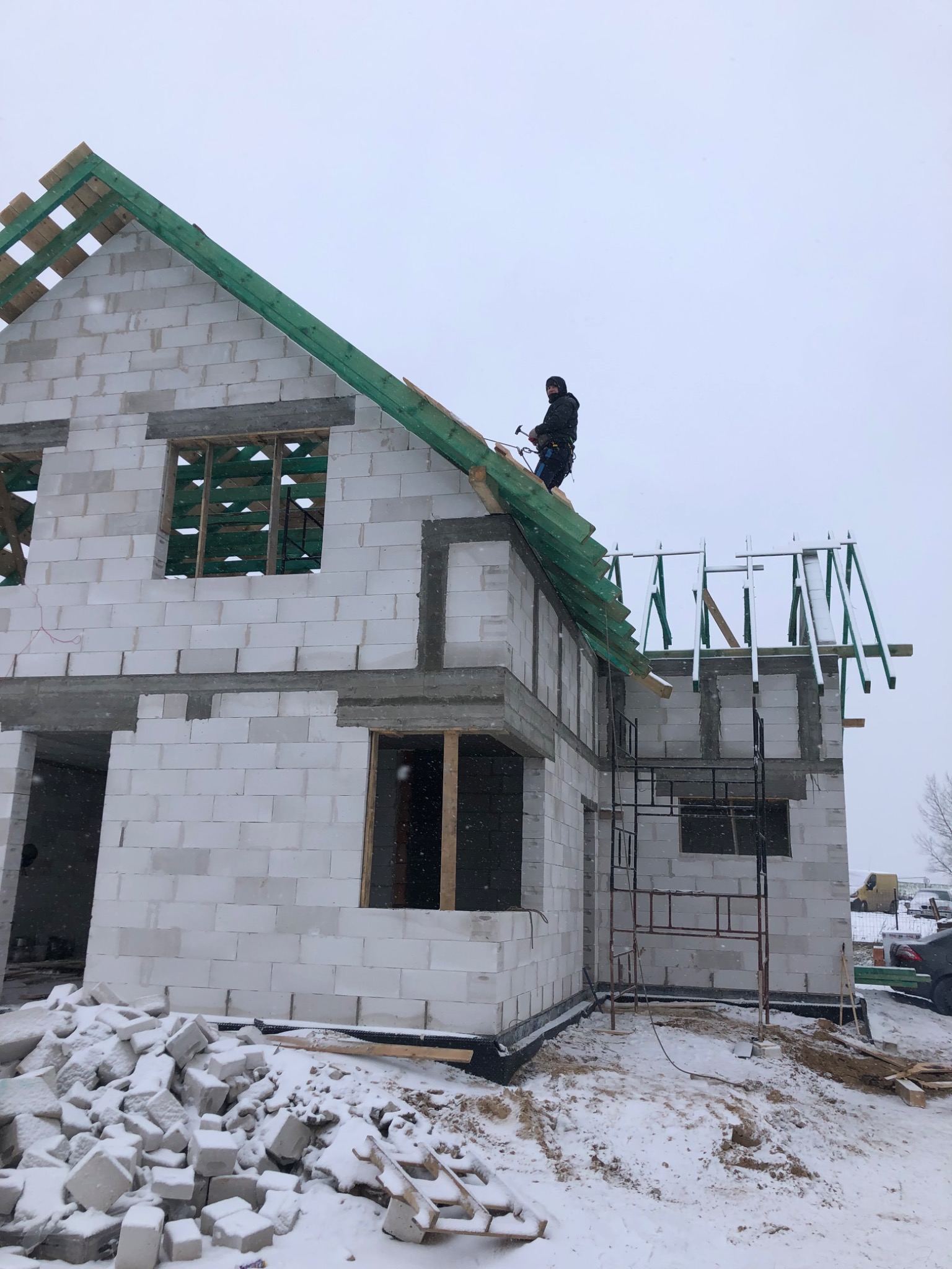 Budowa domu z białych bloczków w zimowej scenerii; widoczna konstrukcja dachu i rusztowanie przy elewacji bocznej; pracownik na dachu z narzędziami.