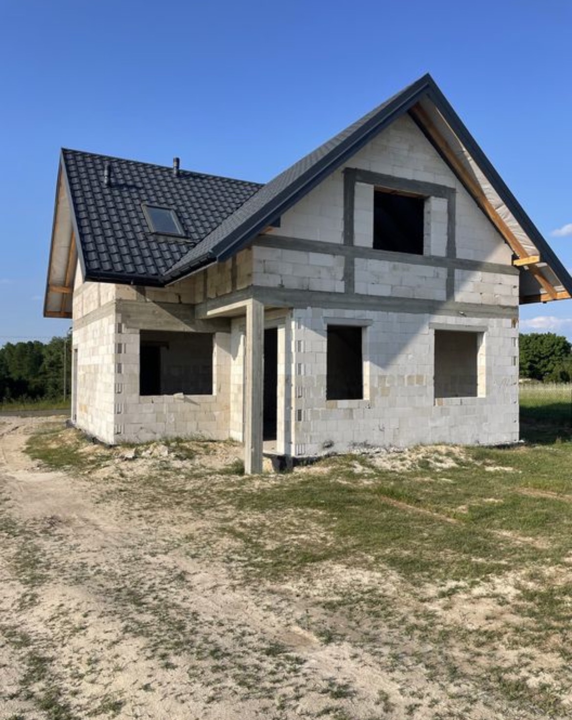 Niedokończony, dwukondygnacyjny dom z pustaków, z ciemną dachówką i oknem dachowym, na działce z piaskiem i trawą, w słoneczny dzień.