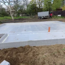 Przygotowany szalunek fundamentowy z izolacją termiczną i wystającą rurą kanalizacyjną na działce budowlanej z przyczepą w tle.