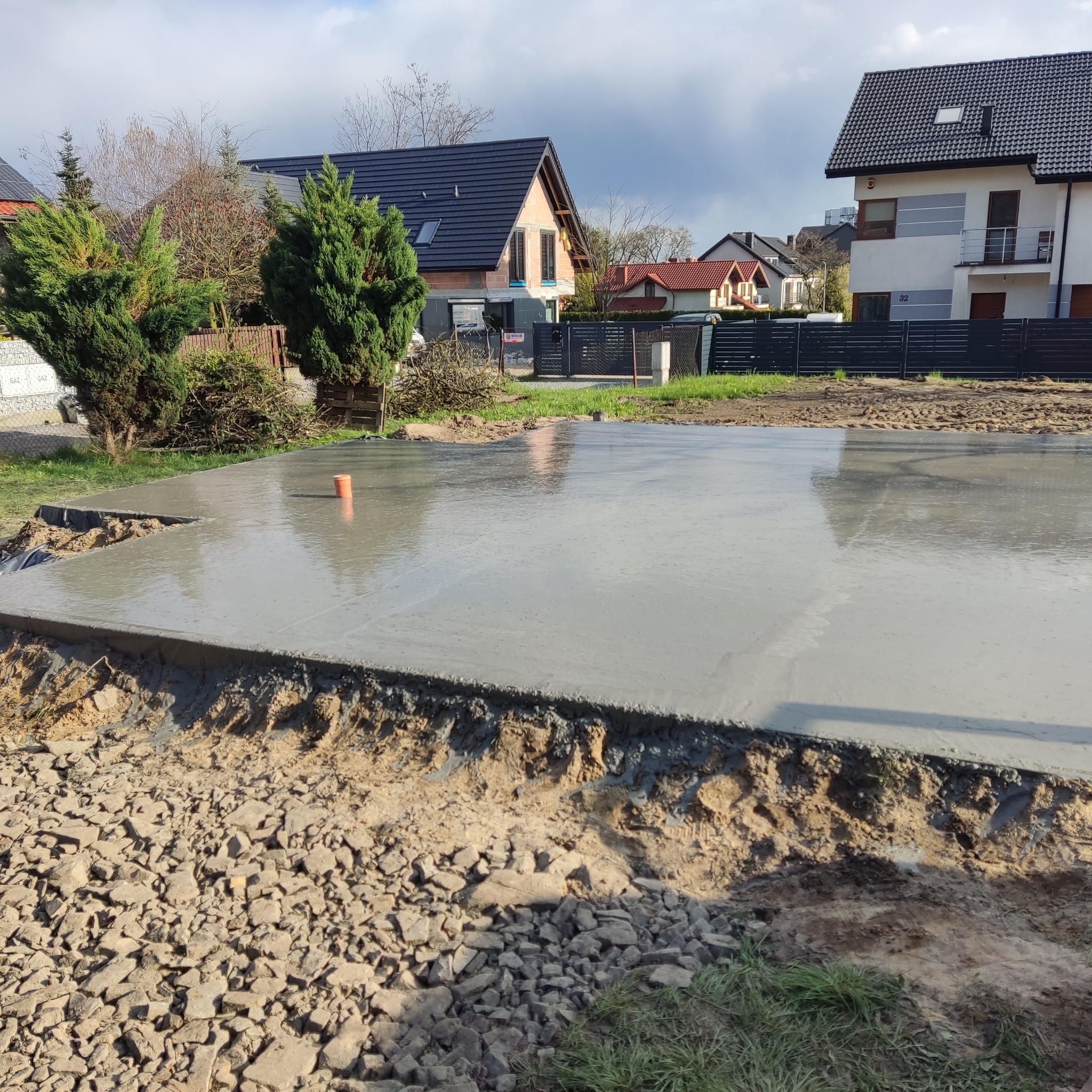 Świeżo wylana, gładka płyta betonowa pod budowę domu jednorodzinnego, z wystającym pomarańczowym rurą w rogu, otoczona ziemią i gruzem, z widocznymi domami w tle.