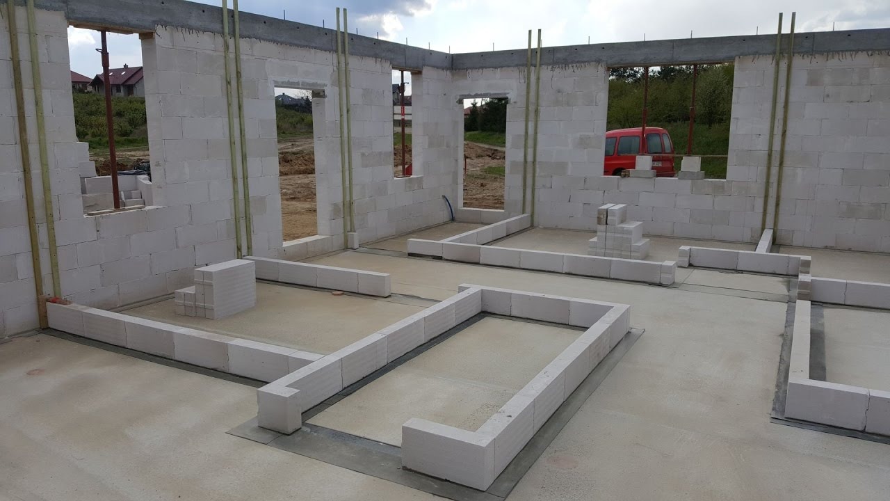 Wnętrze domu w trakcie budowy: wzniesione ściany z białych bloczków, widoczne otwory okienne i drzwiowe, fundamenty pod ściany działowe, czerwony samochód w tle za oknem.