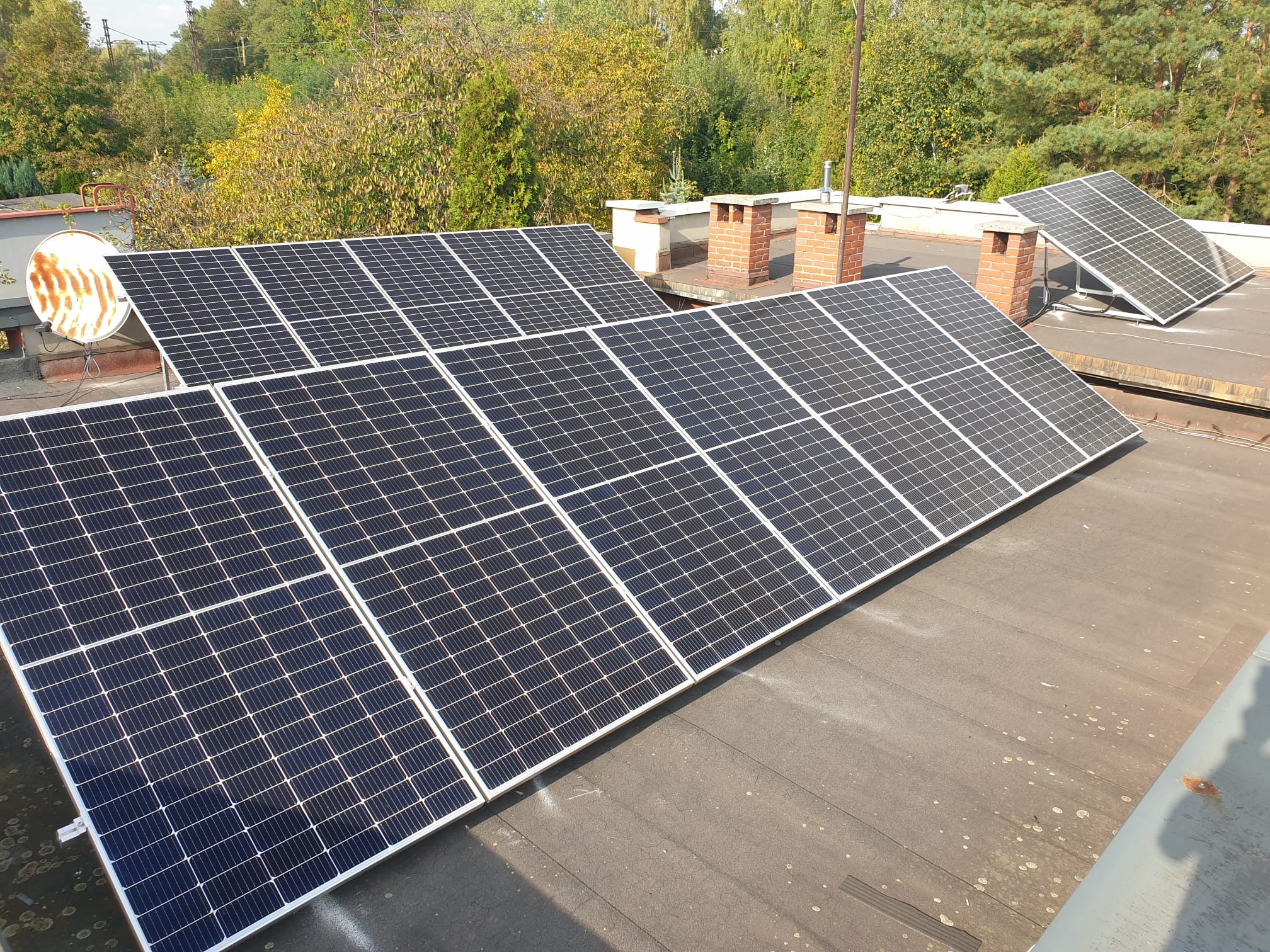 Panele słoneczne na płaskim dachu budynku z kominami w tle, widok z góry.