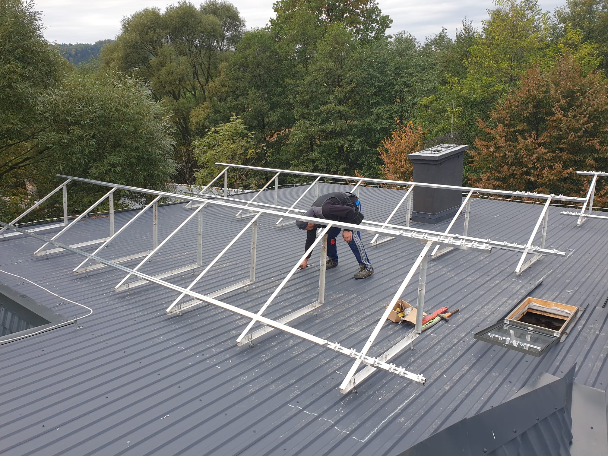 Montaż konstrukcji wsporczej pod panele słoneczne na szarym, blaszanym dachu; widoczne narzędzia i otwarte okno dachowe.
