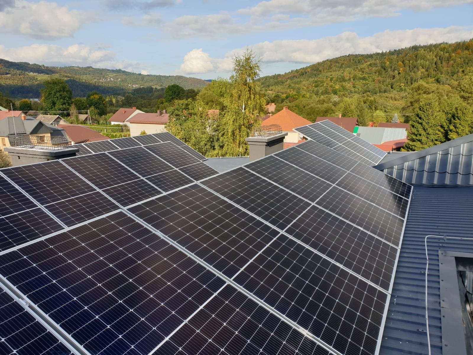 Panele słoneczne zamontowane na dachu domu z widokiem na zalesione wzgórza w tle, w słoneczny dzień.