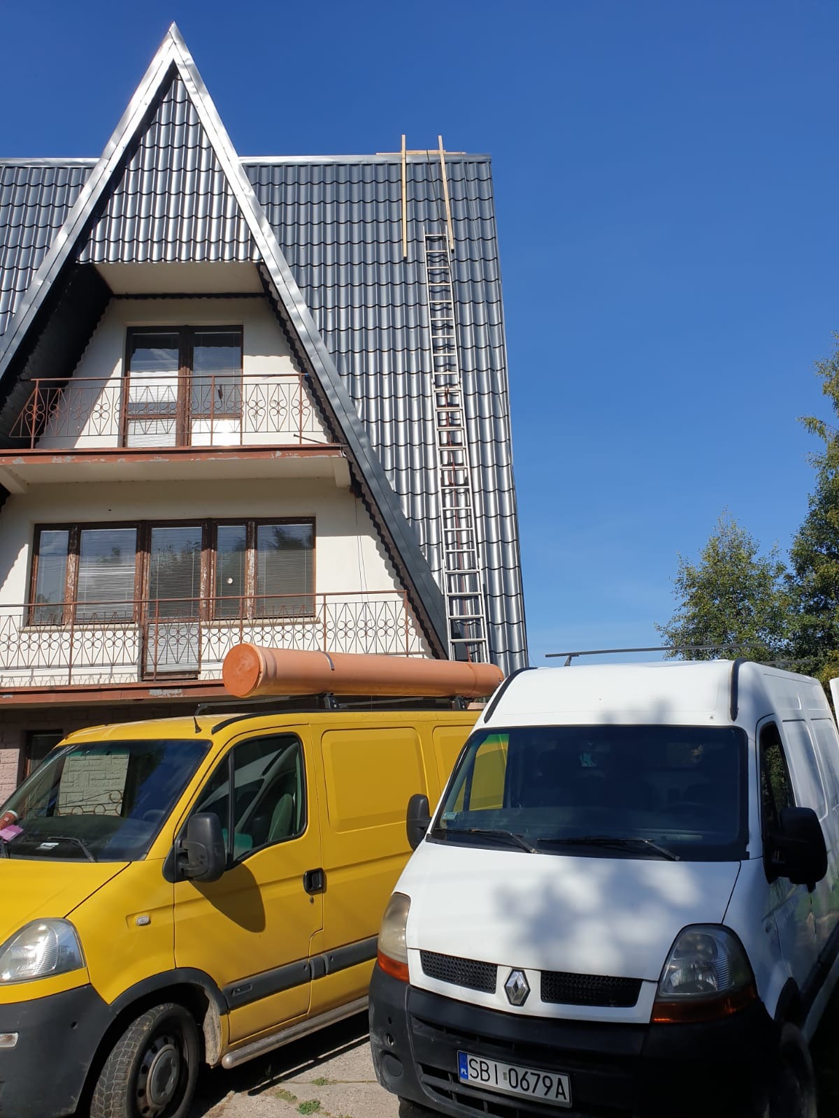 Dwa busy, żółty z rurą na dachu i biały, zaparkowane przed domem z metalowym dachem, na którym oparta jest drabina.