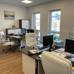 Jasne biuro z kilkoma stanowiskami pracy, widok na zewnątrz przez okna, na biurkach monitory, dokumenty i akcesoria biurowe, na jednym z biurek filiżanka z napojem.