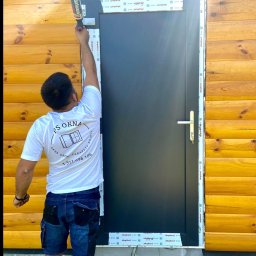 PS OKNA Przemysław Szustek - Montaż drzwi zewnętrznych: monter uszczelnia pianką ramę czarnych drzwi na tle drewnianej elewacji. Widoczny profil Aluplast i narzędzia pracy.