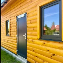 Dom z bali z czarnymi drzwiami i oknami PCV z widocznym odbiciem nieba. Elewacja w ciepłym odcieniu drewna, nowoczesny design, estetyczne wykończenie.