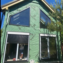 PS OKNA Przemysław Szustek - Budynek w trakcie montażu stolarki okiennej z roletami zewnętrznymi, widoczne okna o nietypowych kształtach i folia paroizolacyjna na ścianach.