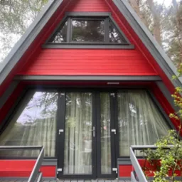 Czerwony domek w stylu A-frame z szarymi detalami, widoczne duże okna i drzwi balkonowe z firanami, otoczony zielenią.