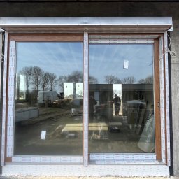 PS OKNA Przemysław Szustek - Brązowe okno z roletą zewnętrzną zamontowane w surowym murze z betonu komórkowego. Widoczne elementy montażowe i folia ochronna.