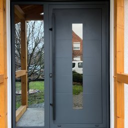PS OKNA Przemysław Szustek - Szare drzwi zewnętrzne z długim uchwytem i szybą, widok z zewnątrz, pod zadaszeniem z drewnianą konstrukcją. W szybie odbija się budynek z roletami.