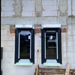 PS OKNA Przemysław Szustek - Dwa okna PCV z roletami zewnętrznymi w surowym murze z betonu komórkowego, widoczne elementy montażowe i rusztowanie w tle, okna z szybami refleksyjnymi.